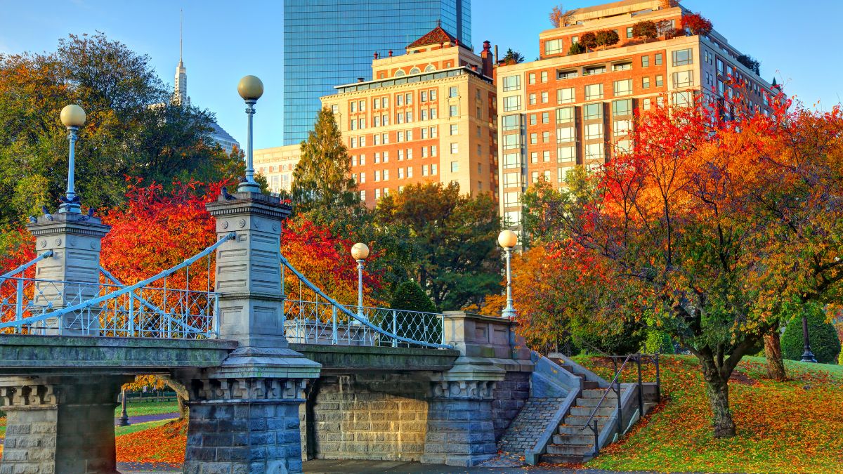 Boston Public Garden bridge framed by red and orange foliage with buildings beyond